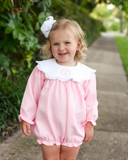 A young toddler girl wearing a pink long-sleeve romper with a large white ruffled collar and a white bow in her hair, standing on a paved path outdoors.