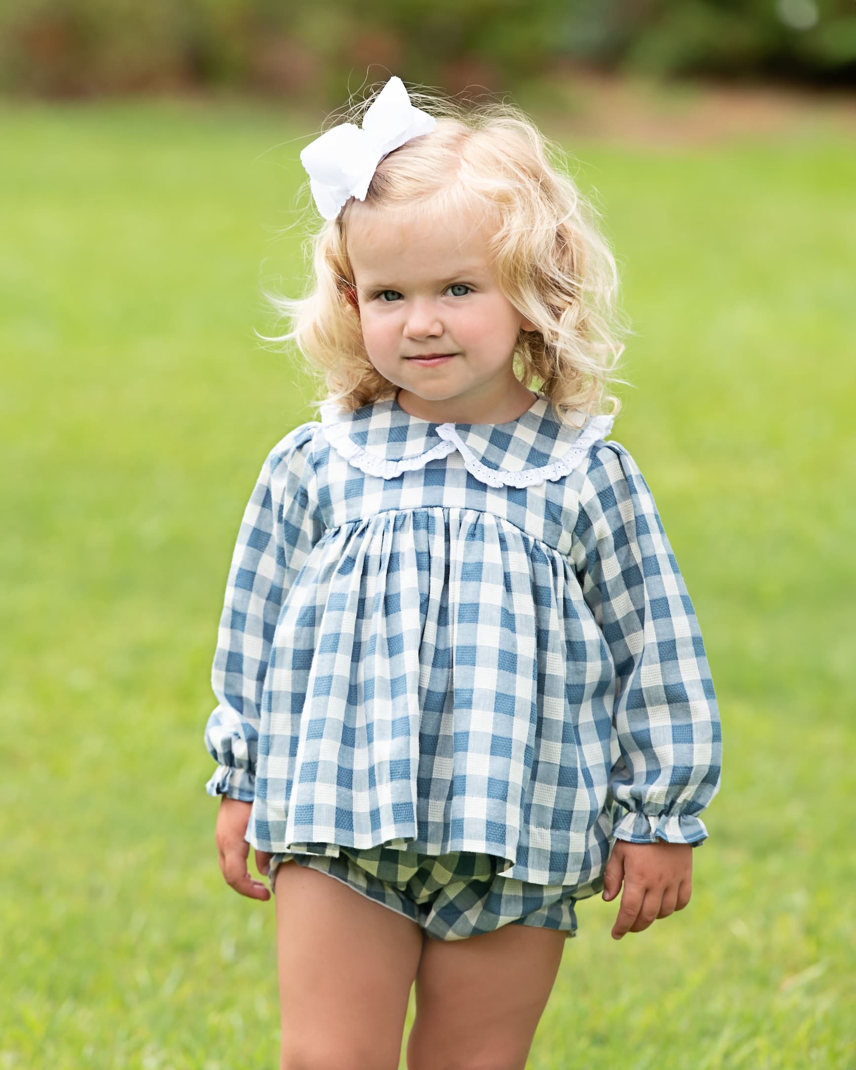 A young toddler girl with blonde curly hair wearing a blue and white gingham long-sleeve top with a white ruffled collar, matching gingham shorts, and a white bow in her hair, standing on a green grassy lawn.