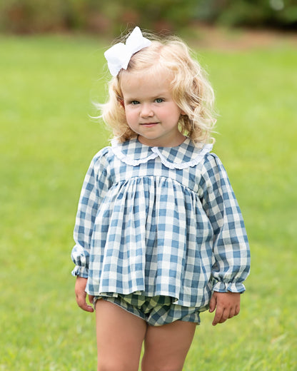 A young toddler girl with blonde curly hair wearing a blue and white gingham long-sleeve top with a white ruffled collar, matching gingham shorts, and a white bow in her hair, standing on a green grassy lawn.