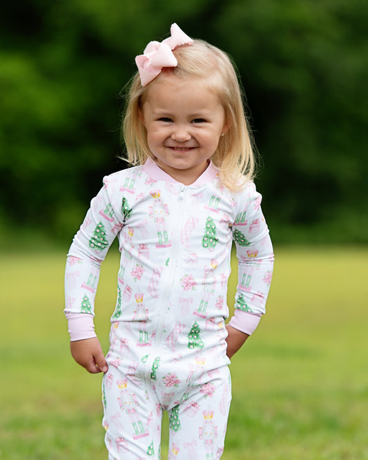 A young toddler girl with blonde hair and a pink bow stands outdoors on a grassy lawn wearing a white long-sleeve pajama top and matching pants with a festive Christmas print of green trees, pink presents, and candy canes.