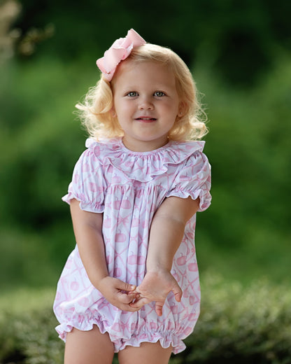 A young toddler girl with blonde hair and a pink bow, wearing a pink and white patterned ruffled romper, standing outdoors against a soft green blurred background.