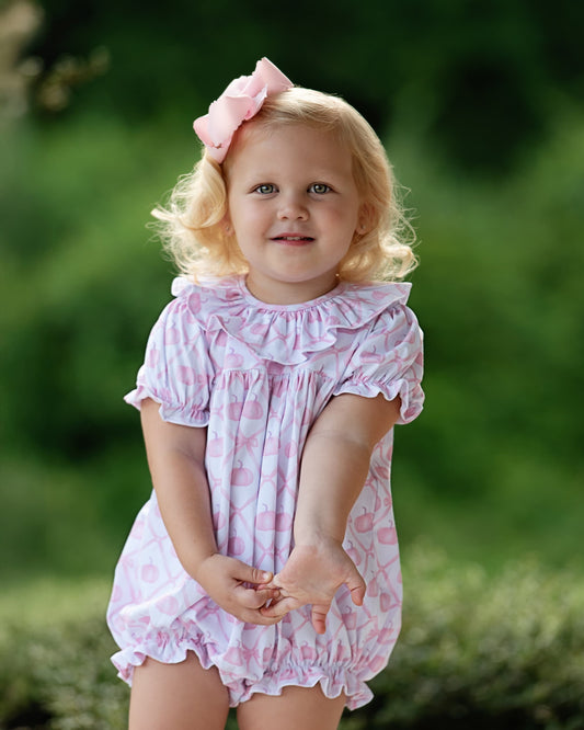 A young toddler girl with blonde hair and a pink bow, wearing a pink and white patterned ruffled romper, standing outdoors against a soft green blurred background.