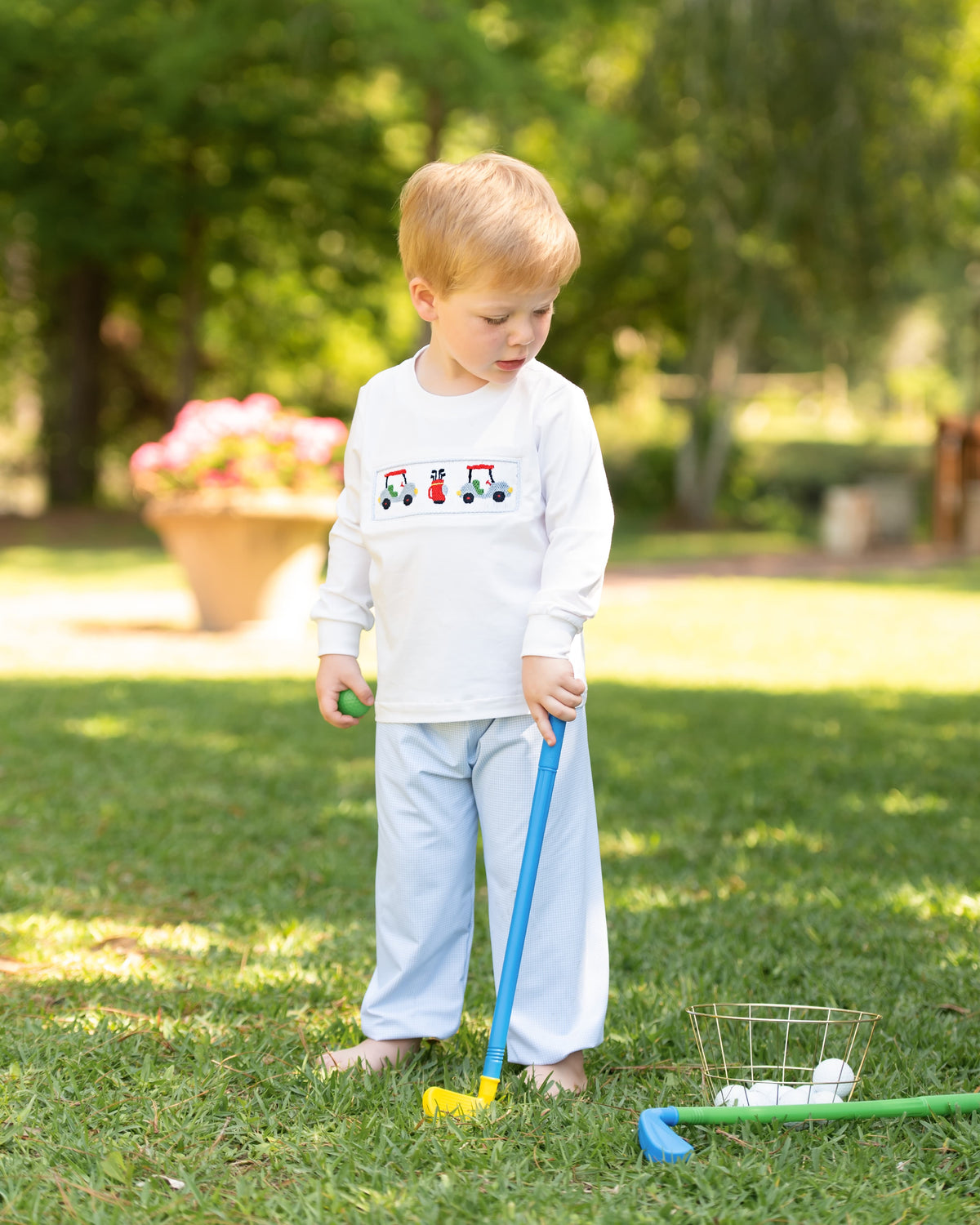 A young toddler stands barefoot on a grassy lawn outdoors, wearing a white long-sleeve shirt with a small tractor graphic on the chest and light blue pants. The child is holding a toy golf club with a yellow head, with golf balls in a wire basket nearby.