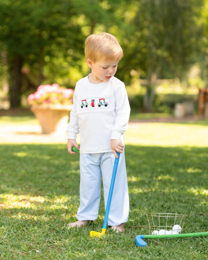 A young toddler stands barefoot on a grassy lawn outdoors, wearing a white long-sleeve shirt with a small tractor graphic on the chest and light blue pants. The child is holding a toy golf club with a yellow head, with golf balls in a wire basket nearby.