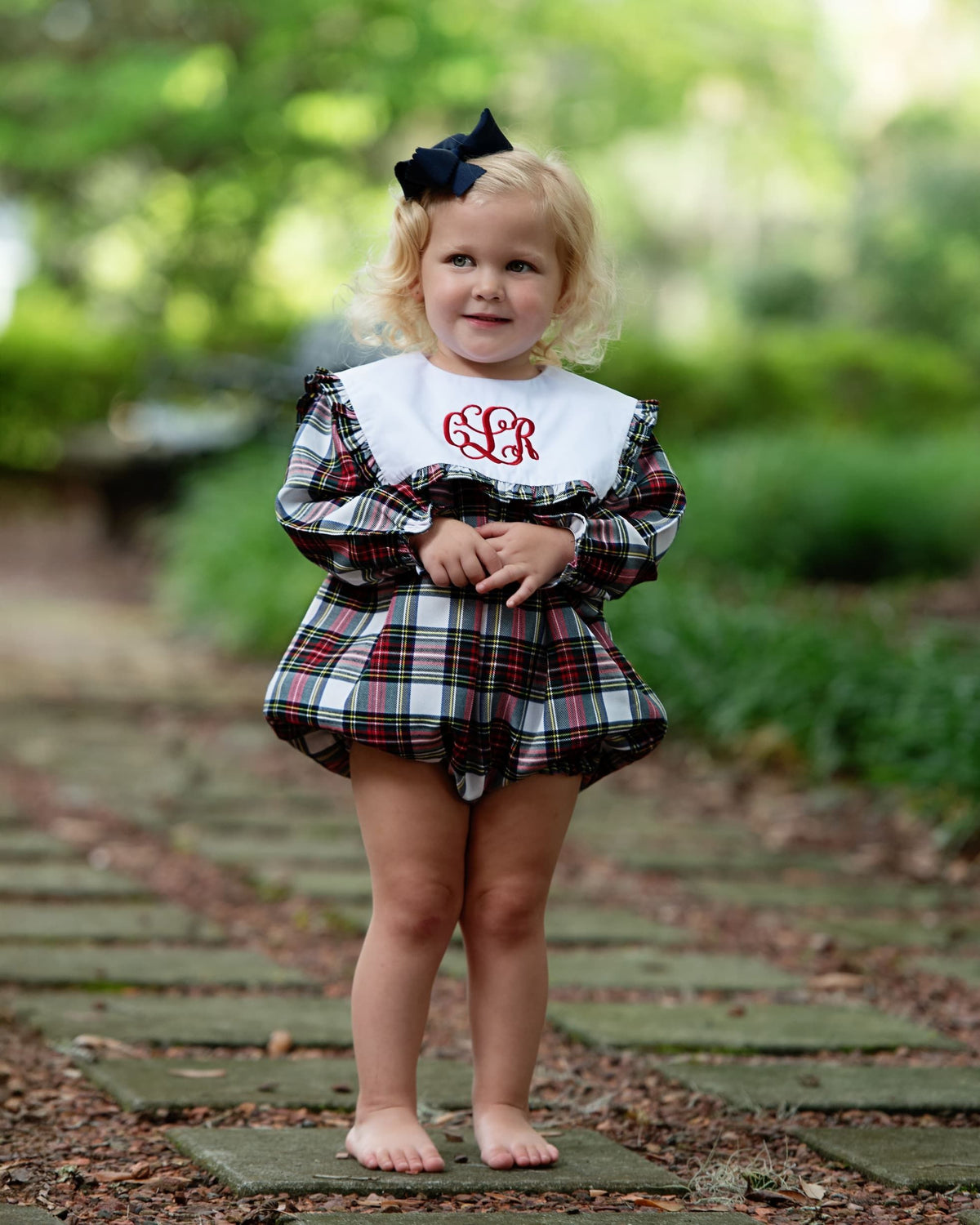 A young toddler stands barefoot on a stone path in a park, wearing a plaid dress with a white bib featuring red monogram lettering.