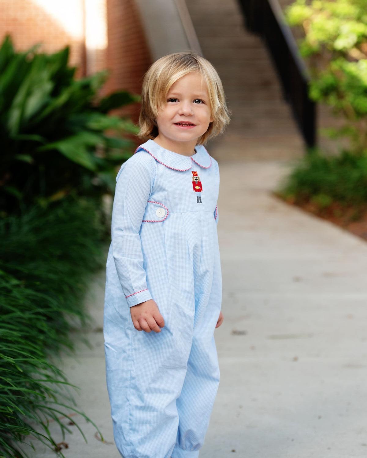 A young toddler with blonde hair stands on an outdoor concrete path wearing a light blue long-sleeve baby jumpsuit with white collar trim and a small red embroidered detail on the chest.