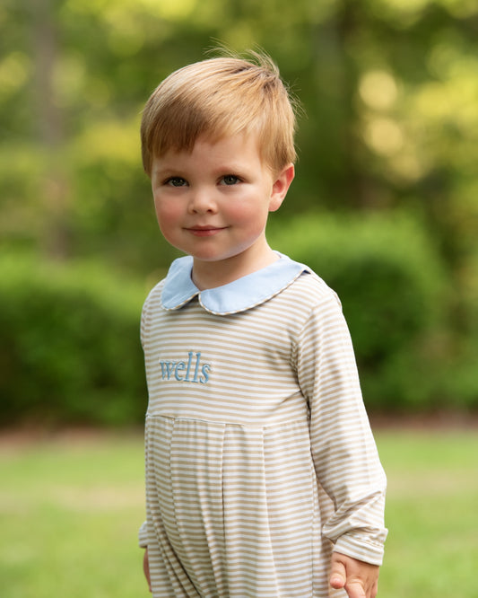 A young toddler with blonde hair stands outdoors in a grassy park setting, wearing a beige and white striped long-sleeve romper with a light blue Peter Pan collar and the name 'wells' embroidered in blue on the chest.