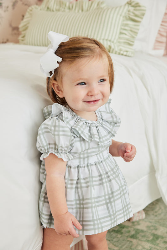 Baby girl in a checkered dress with a white bow in her hair, standing on a bed.