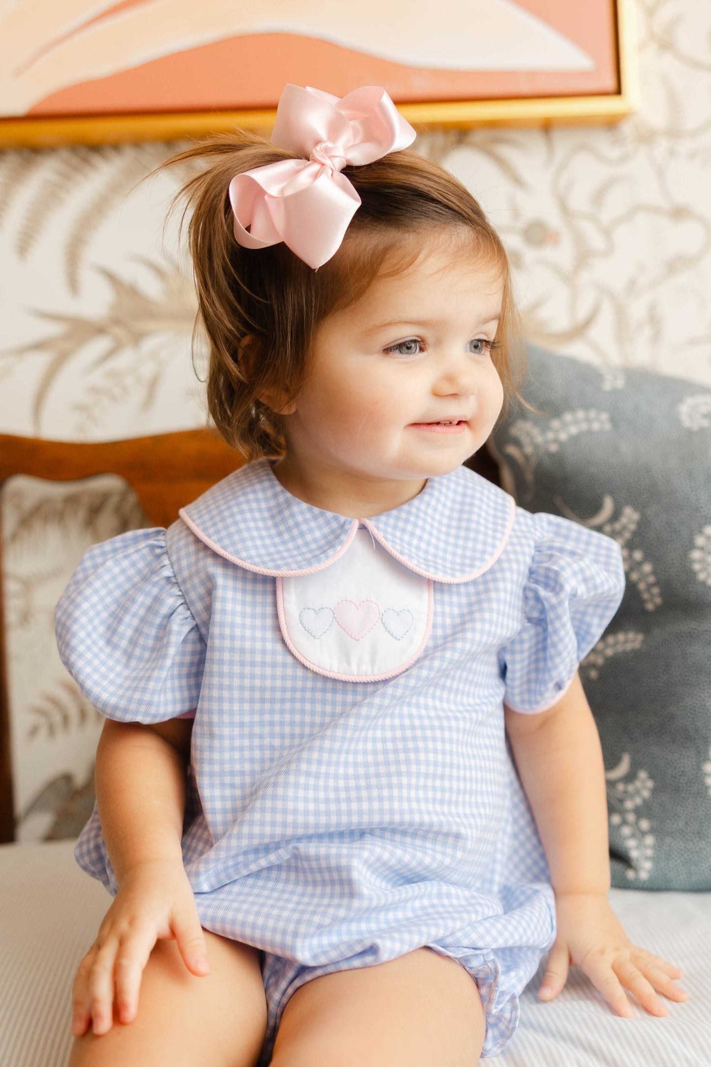 Baby girl wearing a blue checkered outfit with a pink bow in her hair, sitting on a textured surface.