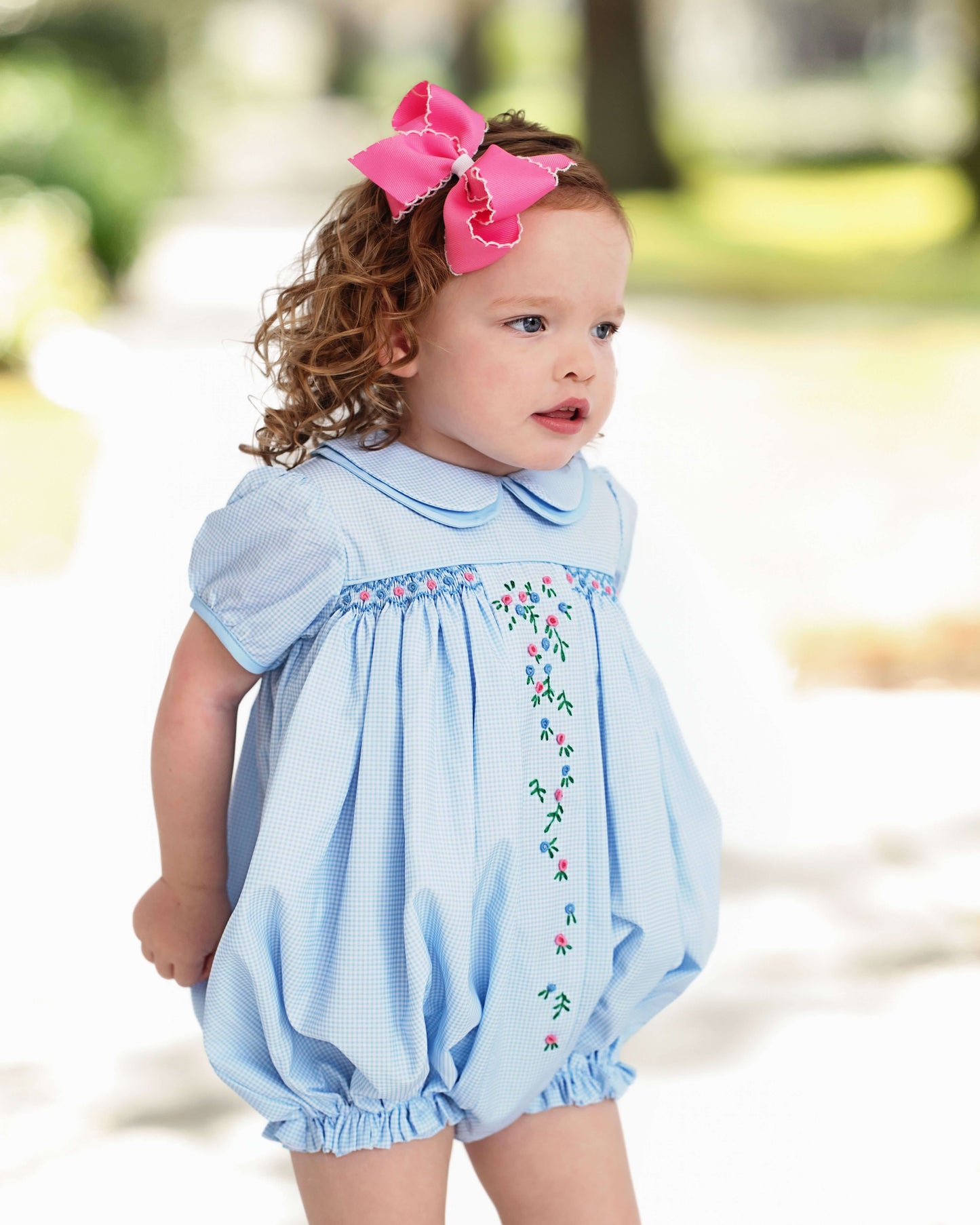 Baby girl wearing a light blue gingham bubble with floral embroidery outdoors.