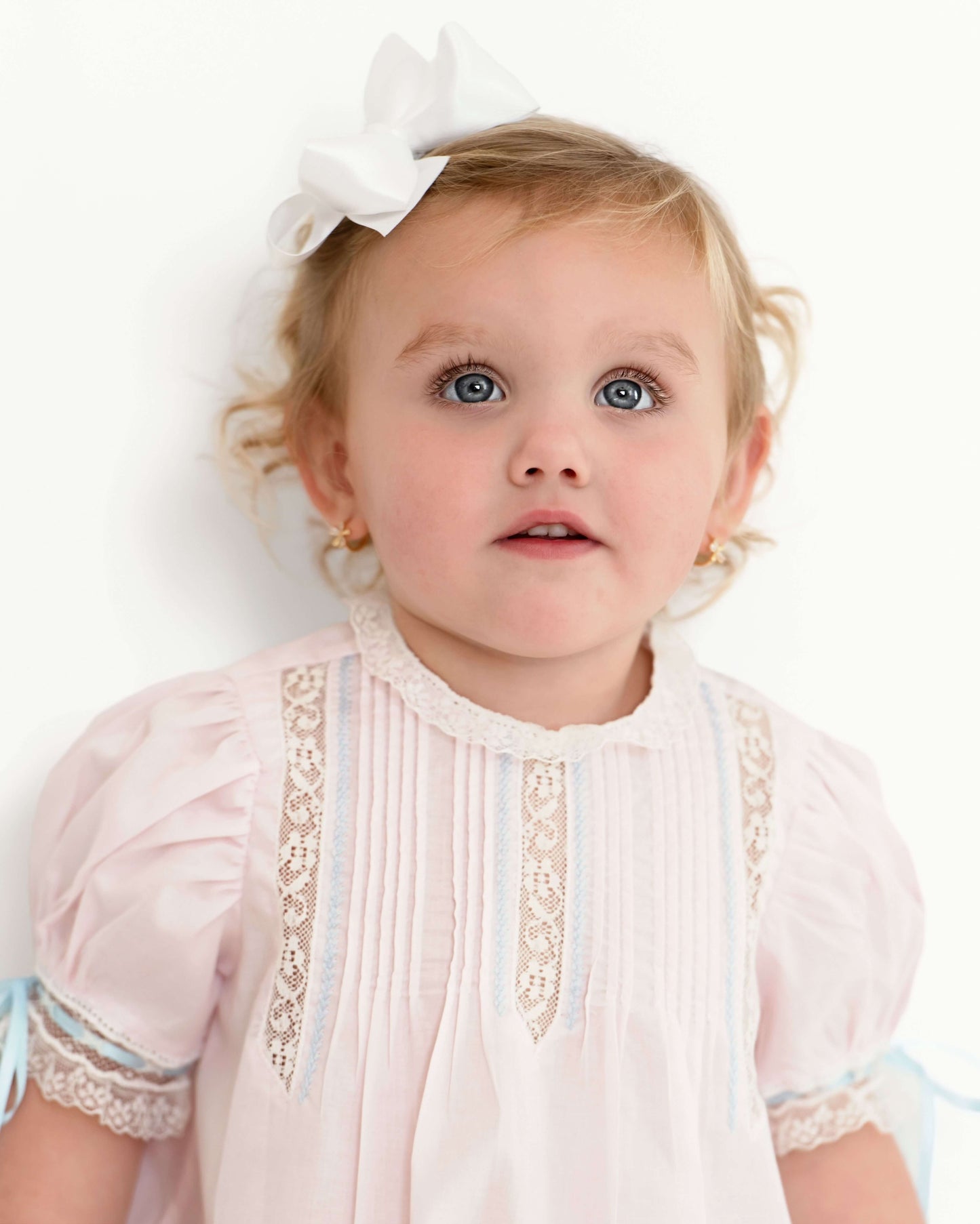 Baby girl wearing a pink dress with lace details and a white bow in her hair.