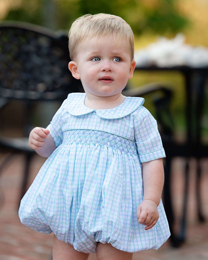 Baby in a blue checkered dress standing outdoors.