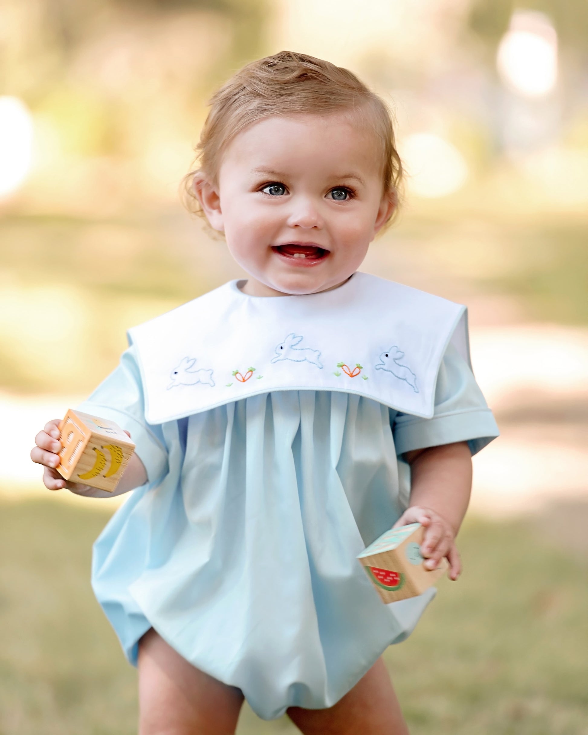 Baby in a light blue bubble with a bib, holding wooden blocks outdoors.