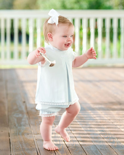 Baby in a light blue dress with a white bow standing on a wooden deck.