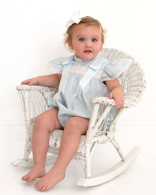 Baby in a light blue dress sitting on a white wicker chair against a white background