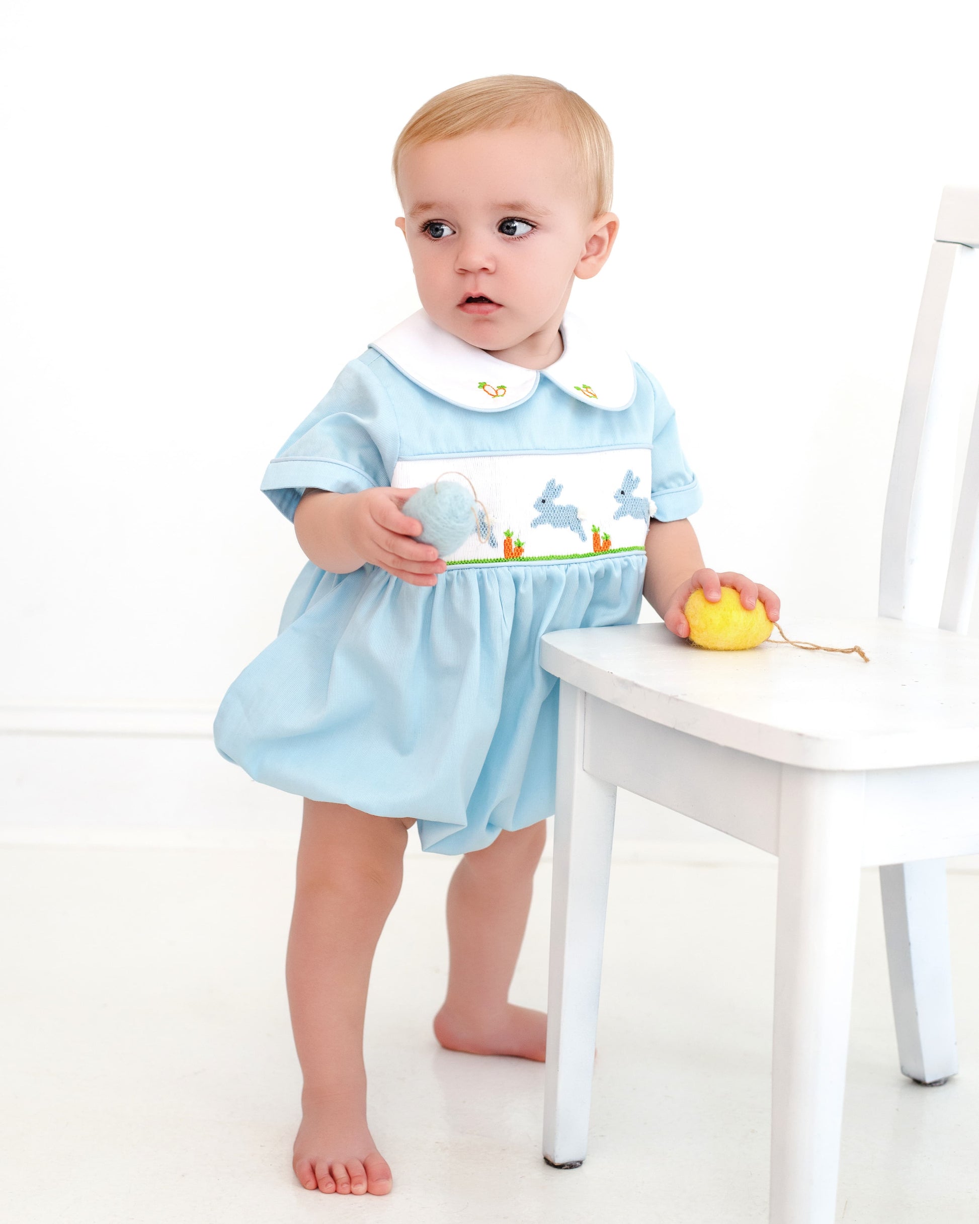 Baby in a light blue dress with a white collar holding a toy, standing next to a white chair on a white background.