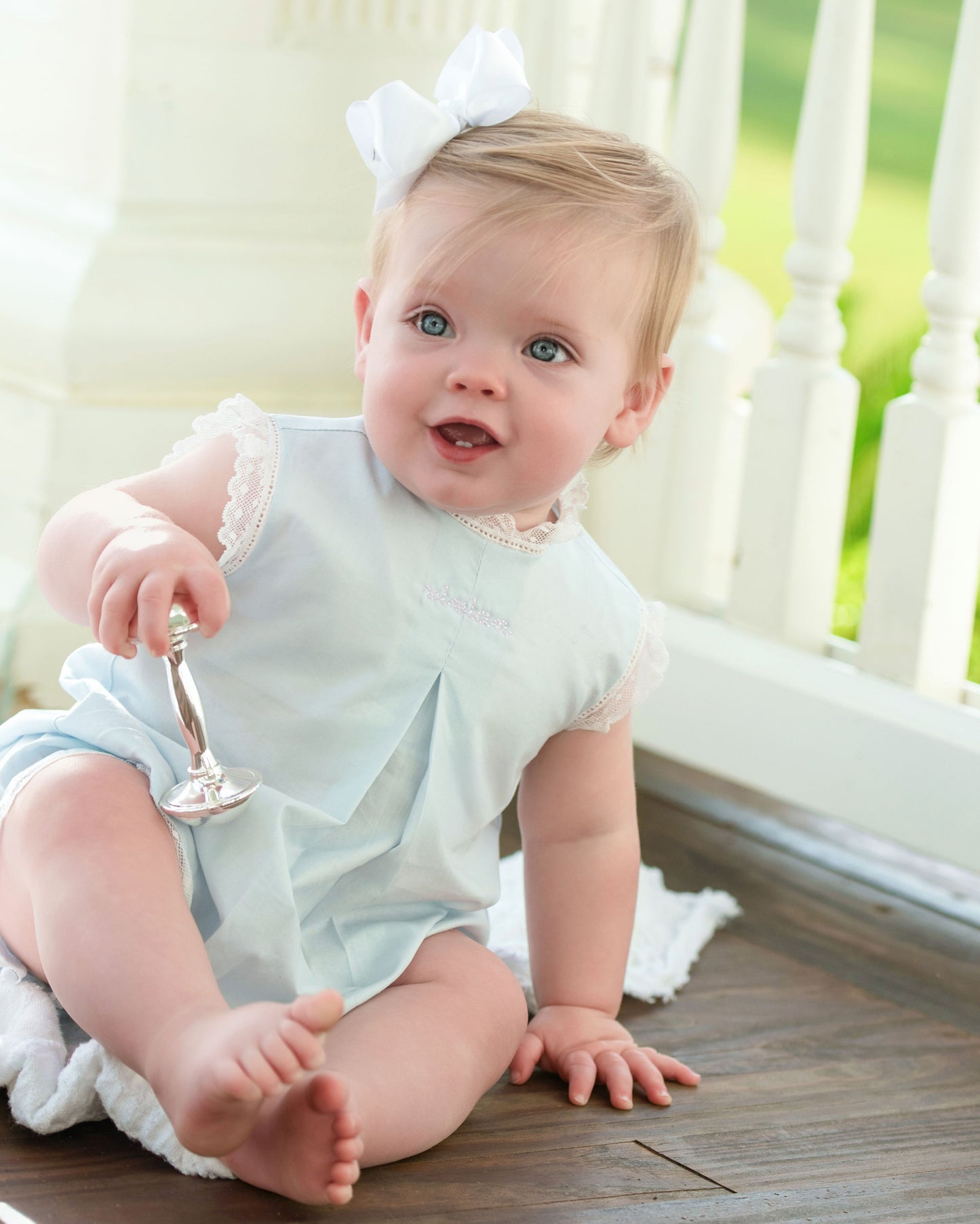 Baby in a light blue outfit with a white bow sitting on a wooden floor.