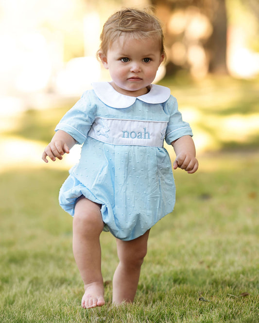 Baby in a light blue romper with a white collar standing on grass.