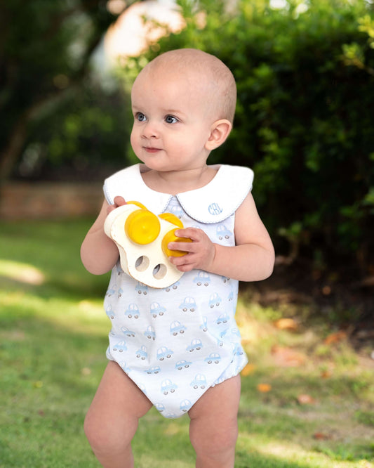 Baby in a white onesie with blue patterns, holding a yellow toy outdoors.