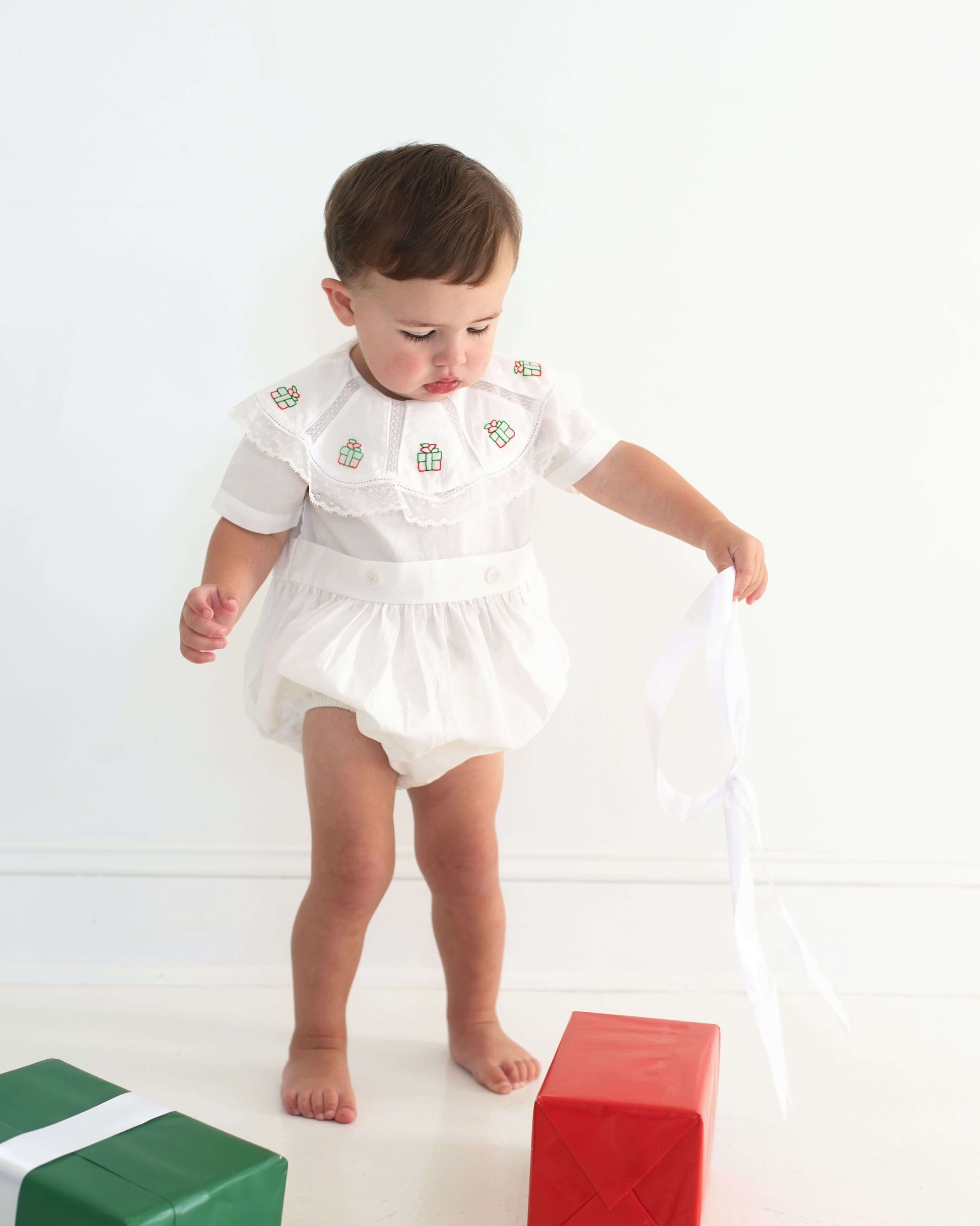 Baby in a white outfit standing on a white background with colorful blocks.