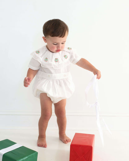 Baby in a white outfit standing on a white background with colorful blocks.