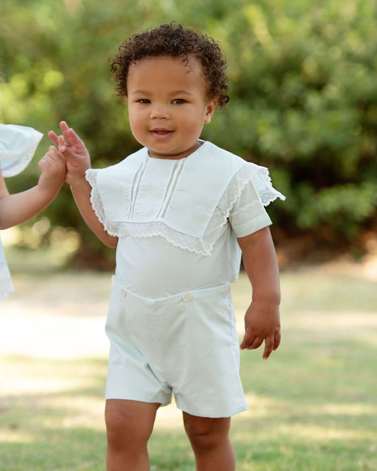 Baby in a white outfit standing outdoors with greenery in the background
