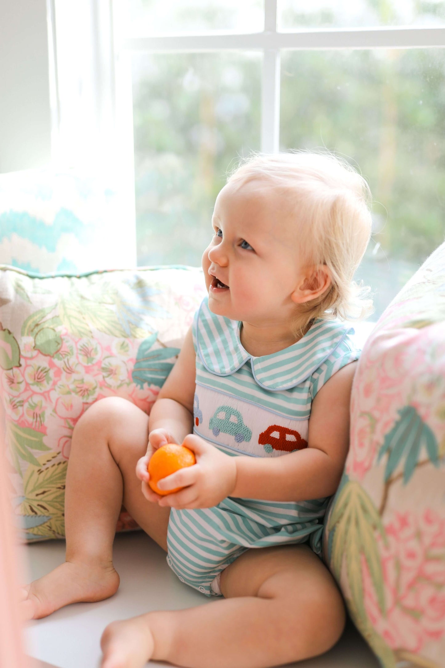 Baby sitting on a couch holding an orange toy, looking out a window with a floral cushion in the background.
