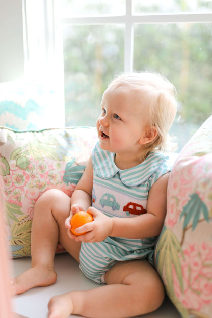 Baby sitting on a couch holding an orange toy, looking out a window with a floral cushion in the background.