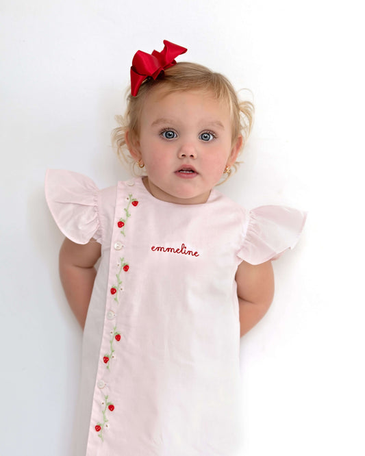 Baby wearing a pink dress with red accents and a red bow on a white background