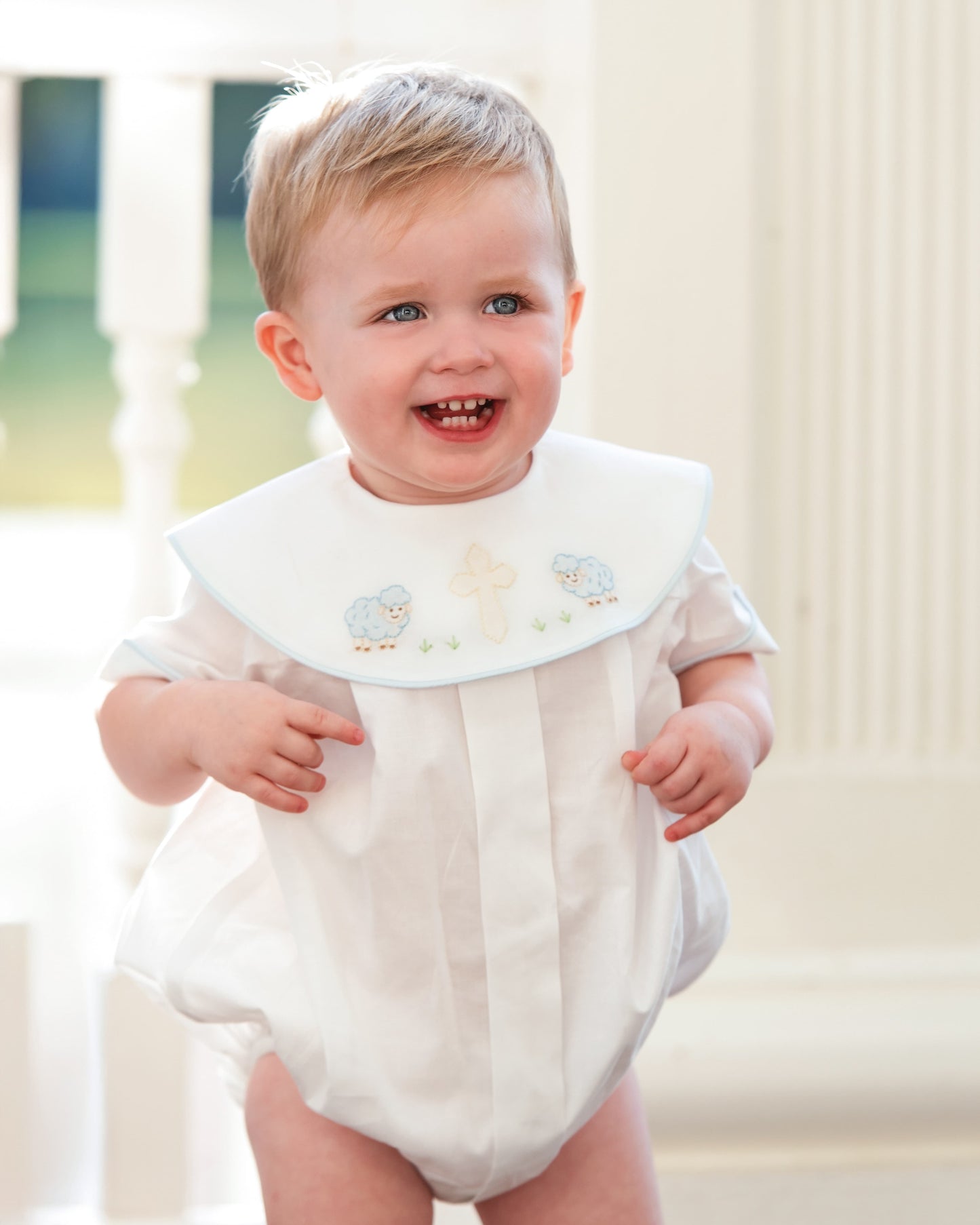 Baby wearing a white outfit with a decorative collar in a softly blurred indoor setting