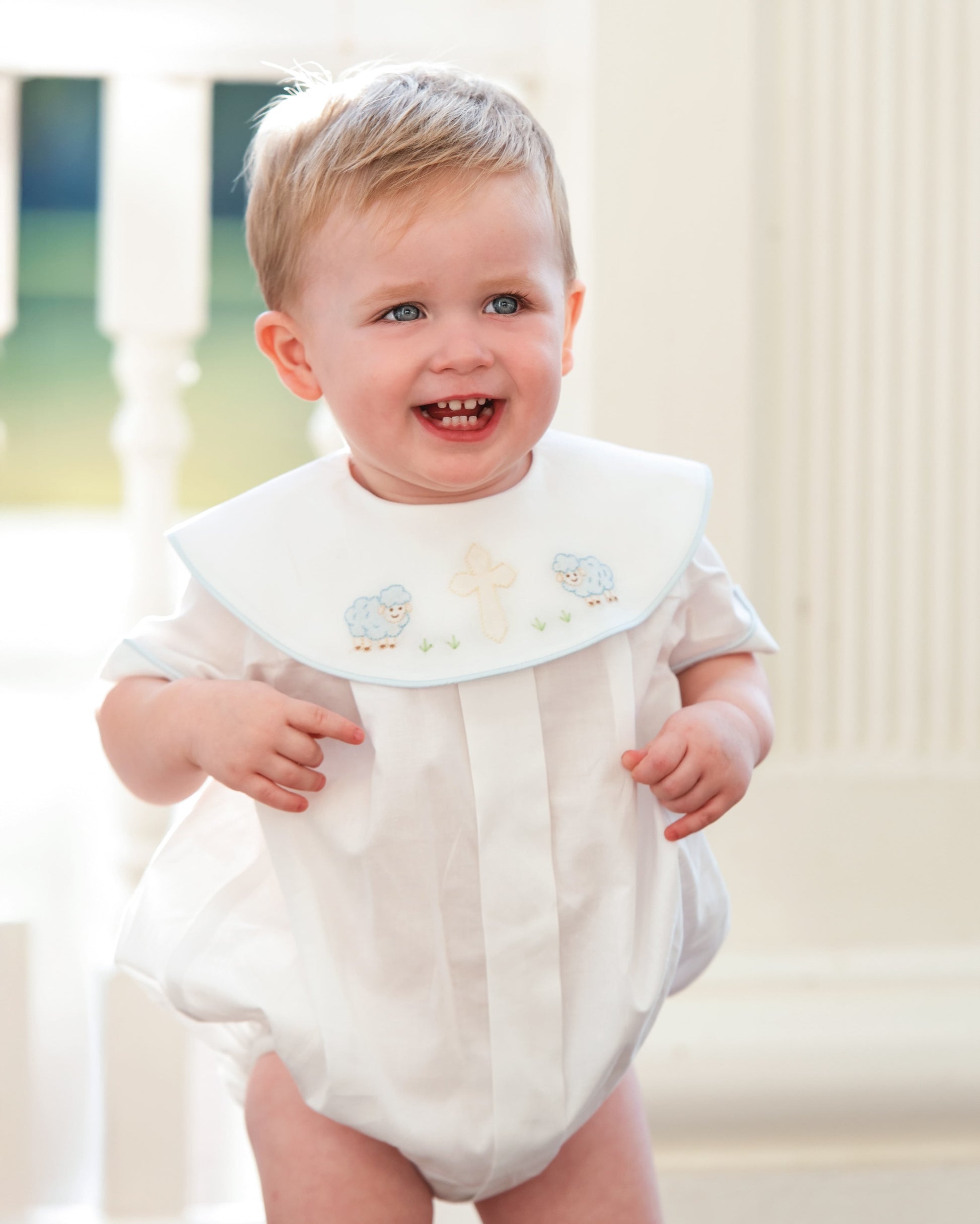 Baby wearing a white outfit with a decorative collar in a softly blurred indoor setting