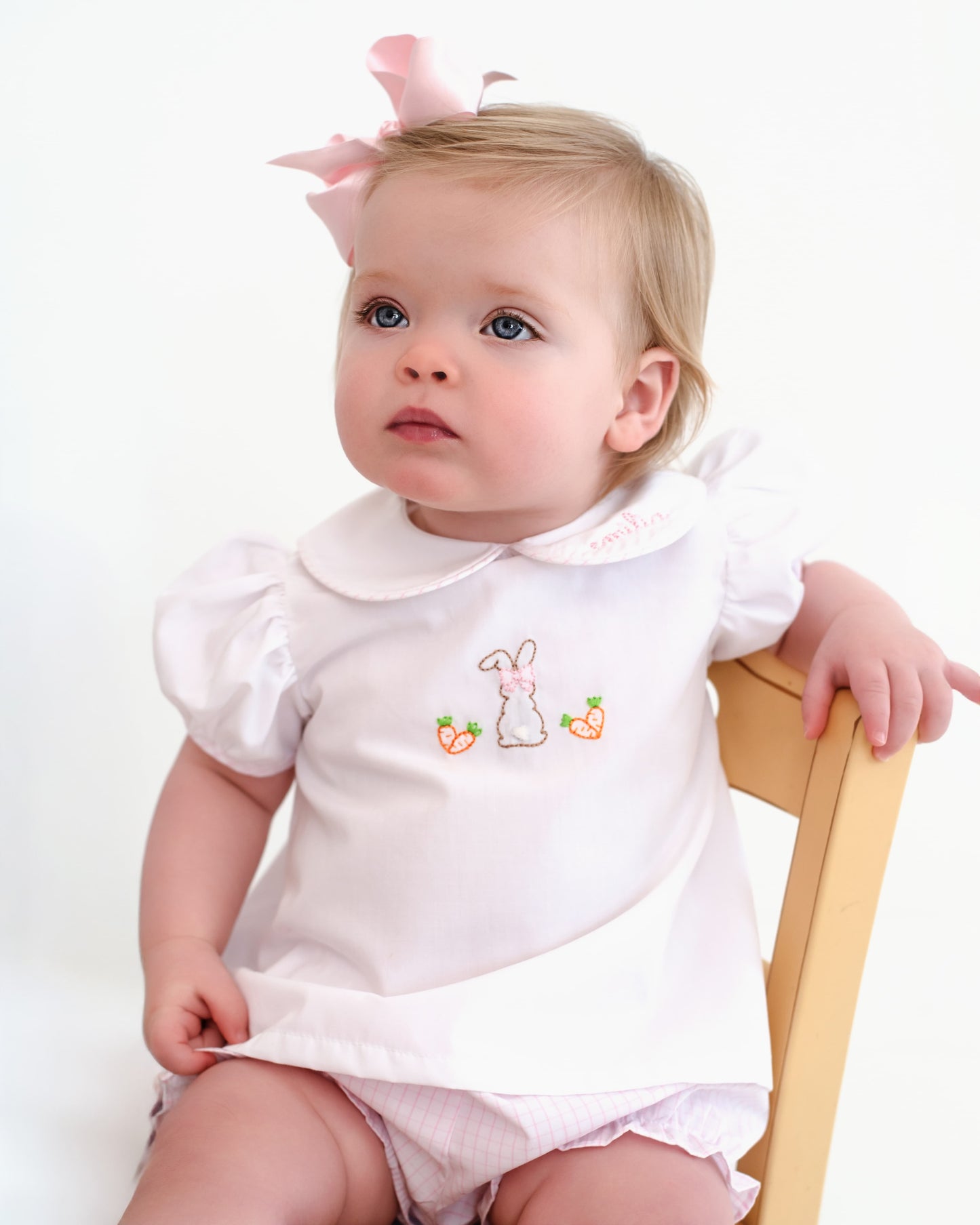 Baby wearing a white outfit with bunny design, sitting on a chair against a white background