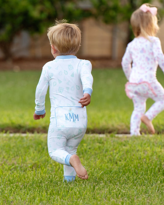 Back view of a toddler boy wearing blue and green birthday print zipper pajamas with a monogrammed flap and light blue trim, running barefoot on the grass next to a girl in matching pink pajamas.