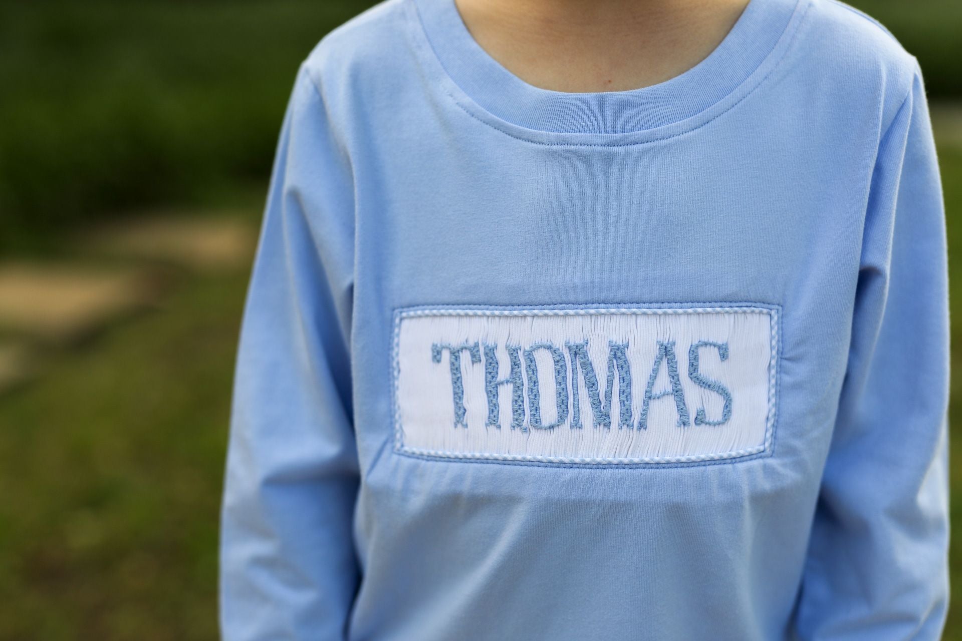 Blue long-sleeve shirt with 'THOMAS' embroidered on a blurred green background
