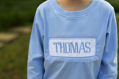 Blue long-sleeve shirt with 'THOMAS' embroidered on a blurred green background