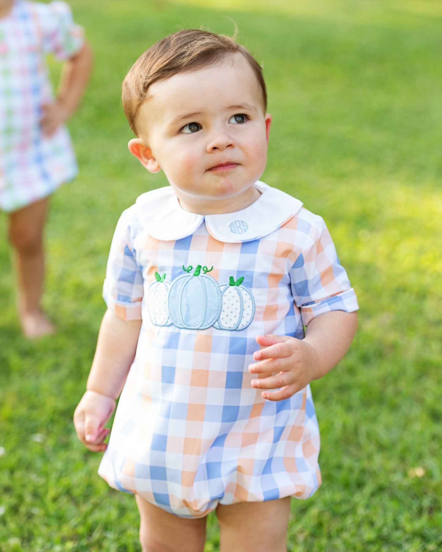 Boy in a checkered romper with pumpkin design standing on grass
