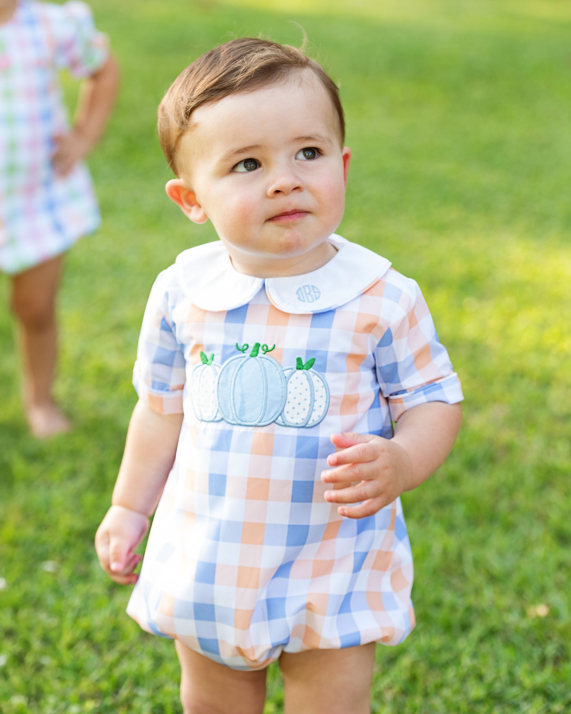 Boy in a checkered romper with pumpkin design standing on grass