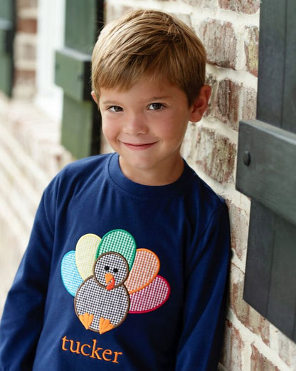 Boy wearing a navy shirt with a colorful turkey design and 'tucker' monogram, standing against a brick wall.