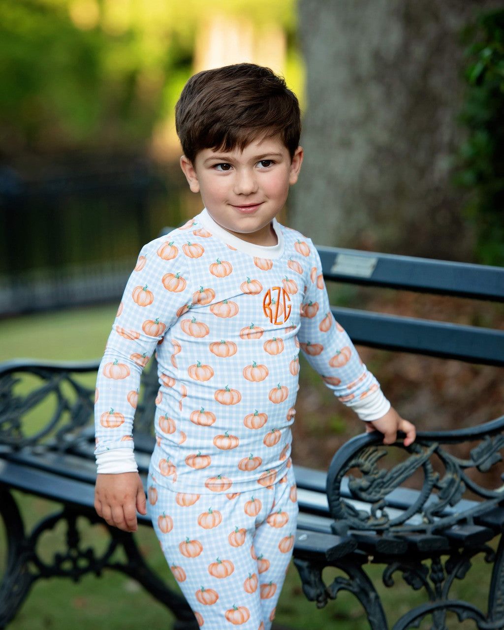 Boy wearing a pumpkin-patterned pajama set standing on a bench outdoors.