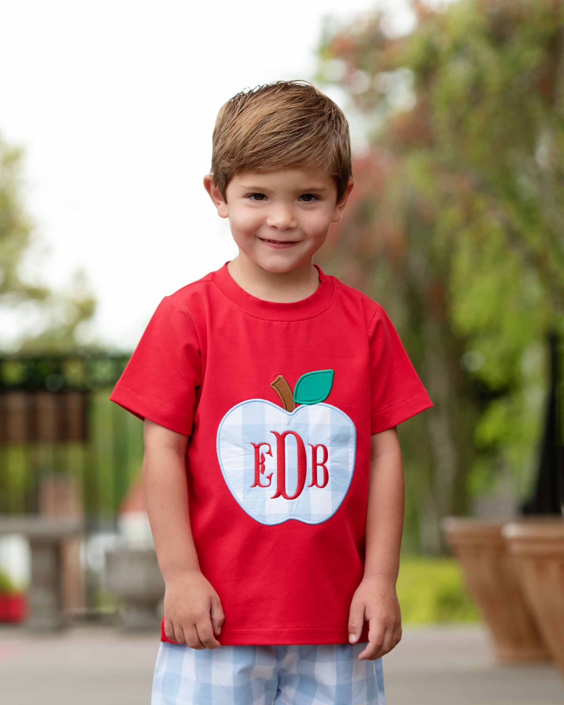 Boy wearing a red shirt with a big apple and blue shorts.