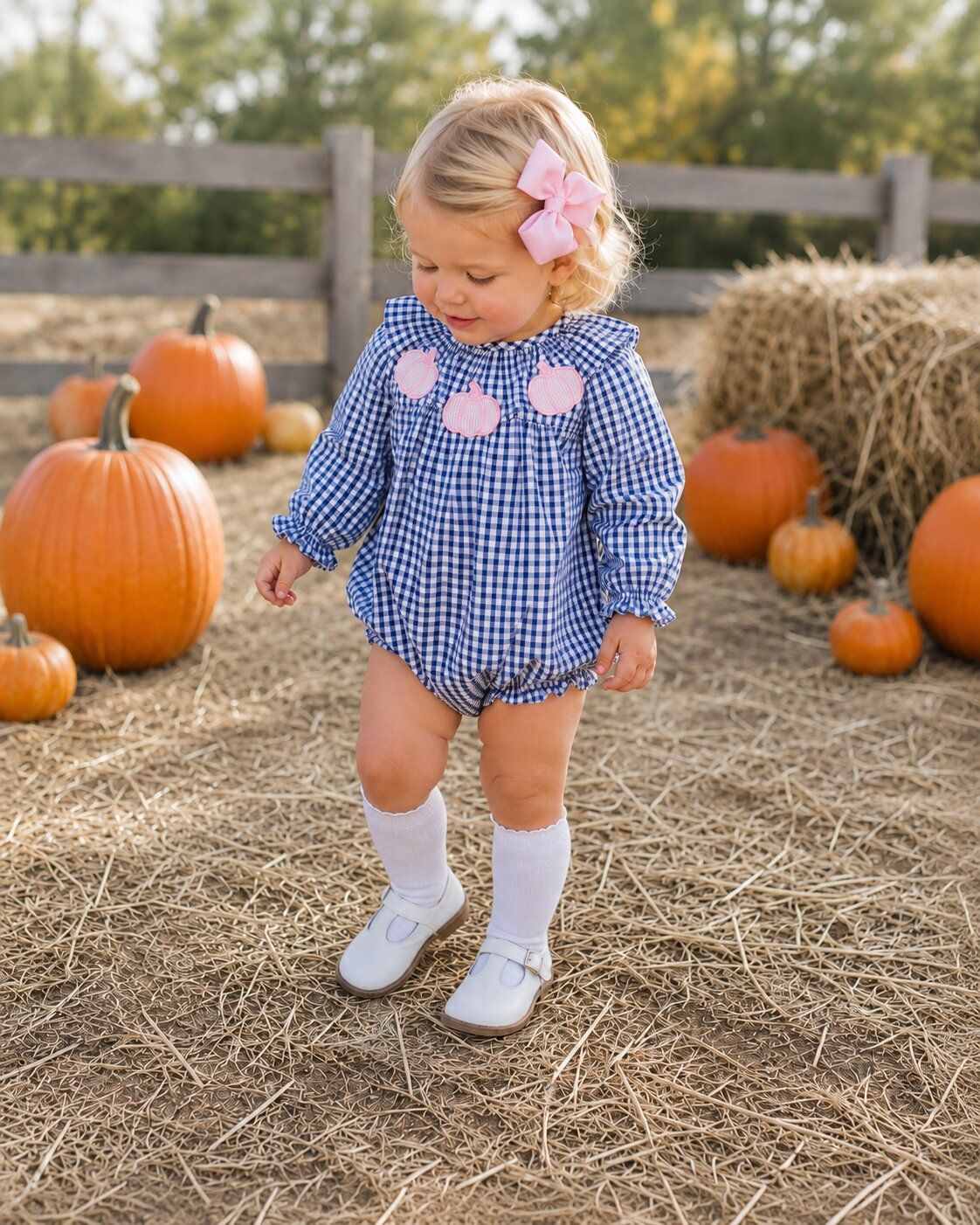 Child in a blue checkered outfit with pink polka dots standing in a pumpkin patch.