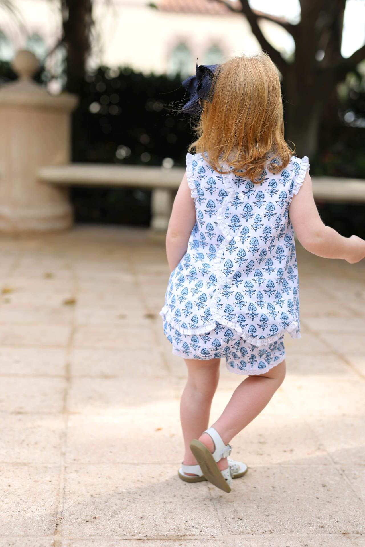 Child in a blue floral outfit walking outdoors on a sunny day.