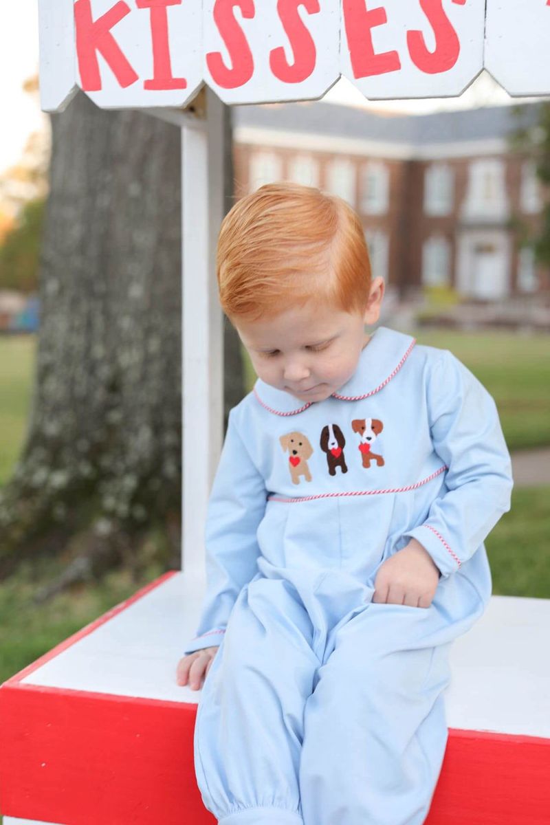 Child in a light blue outfit with puppy designs sitting on a red surface, with 'Kisses' sign in the background.