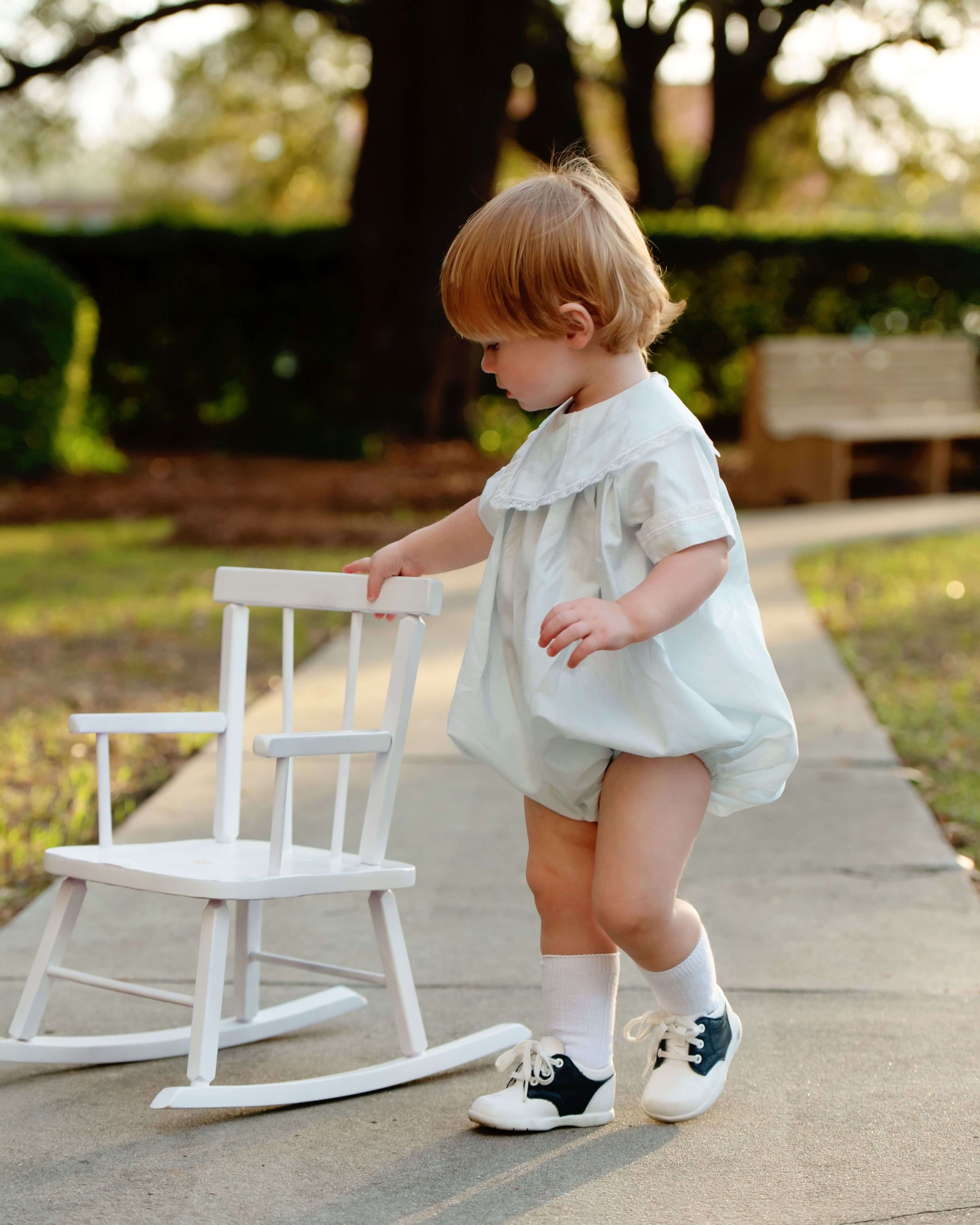 Child in a light dress standing next to a white rocking chair outdoors.