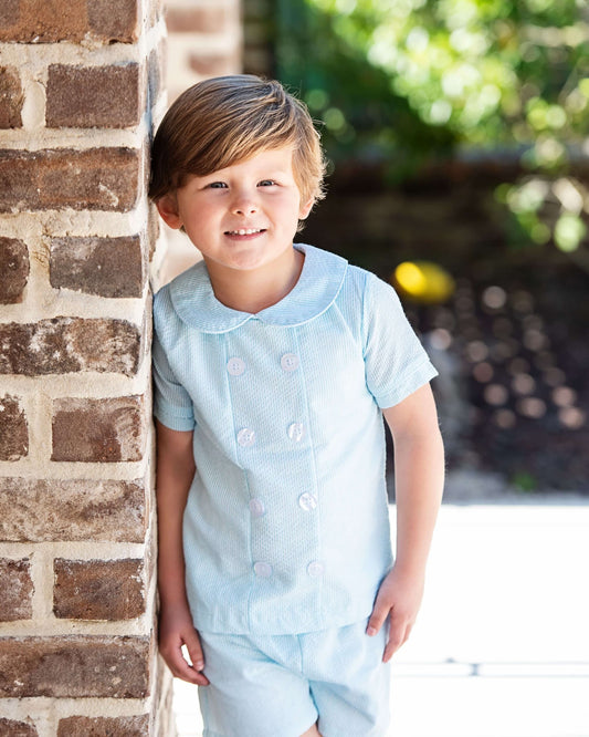 Child in a light blue outfit standing against a brick wall with a blurred outdoor background