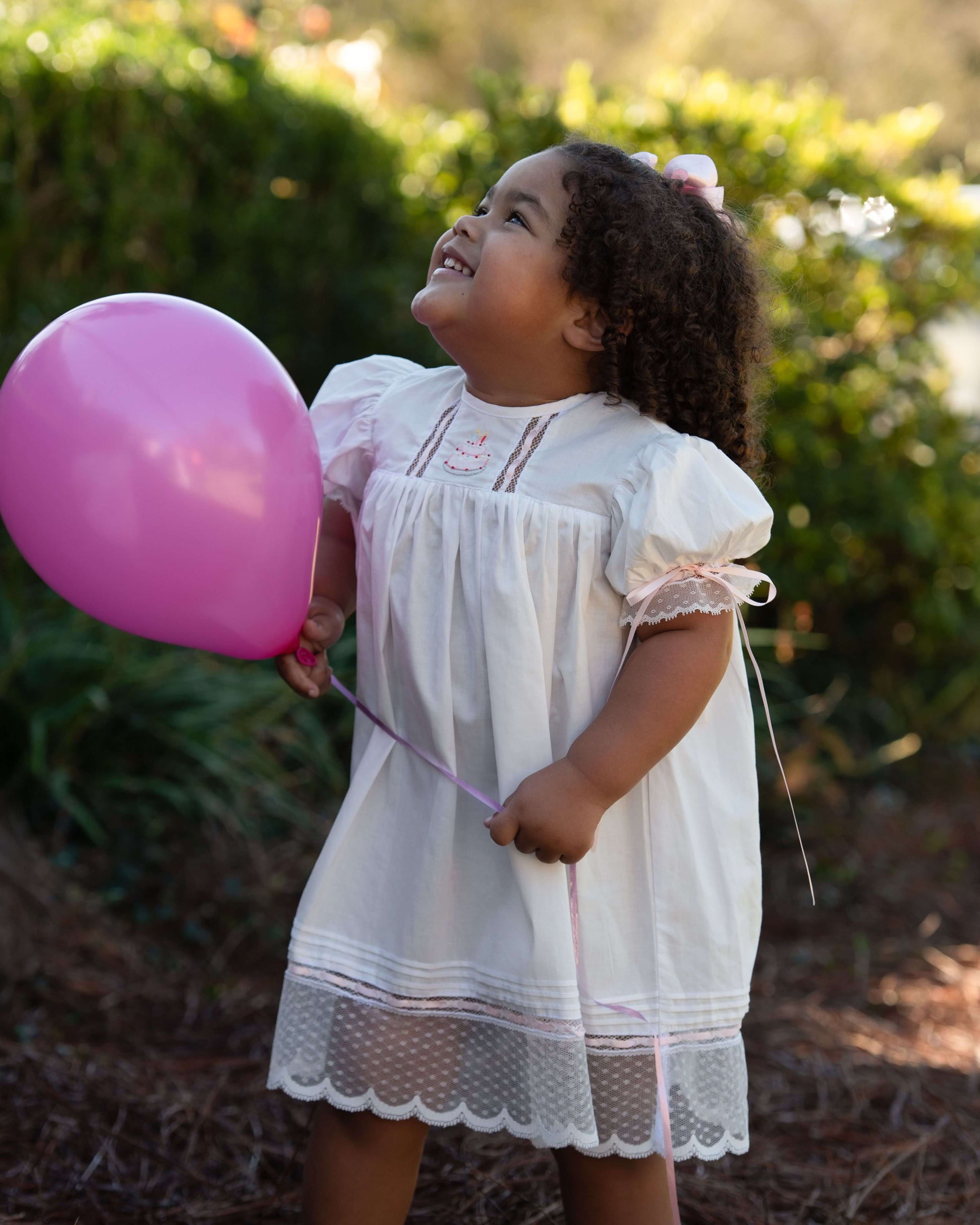 Child in a white dress holding a pink balloon outdoors
