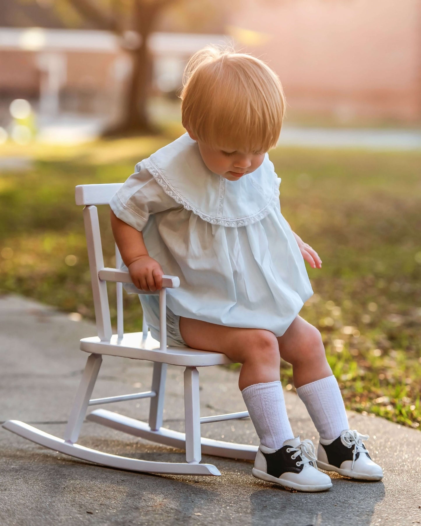 Child in a white dress sitting on a small white rocking chair outdoors.
