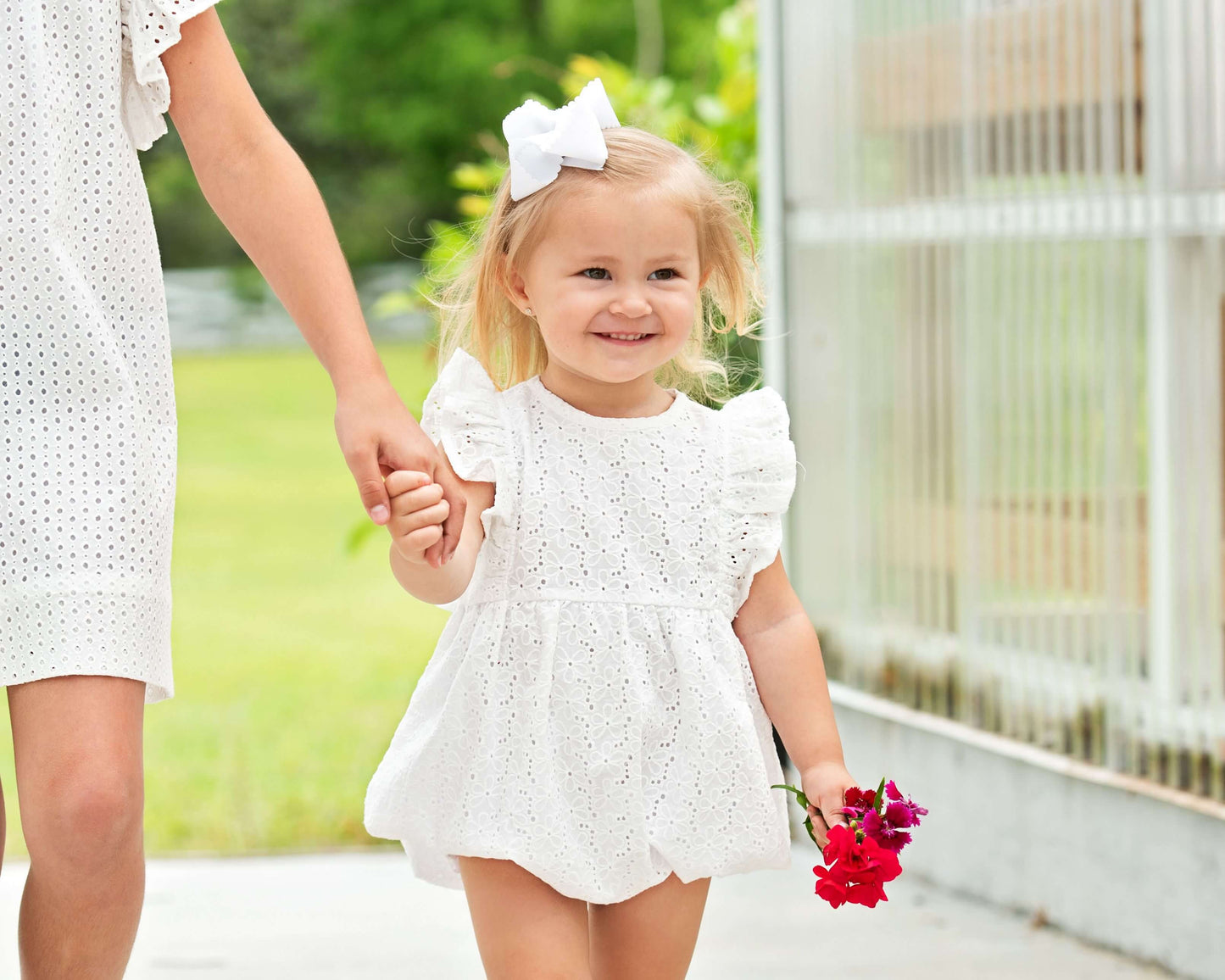 Child in a white eyelet bubble holding hands with a little girl, standing outdoors.