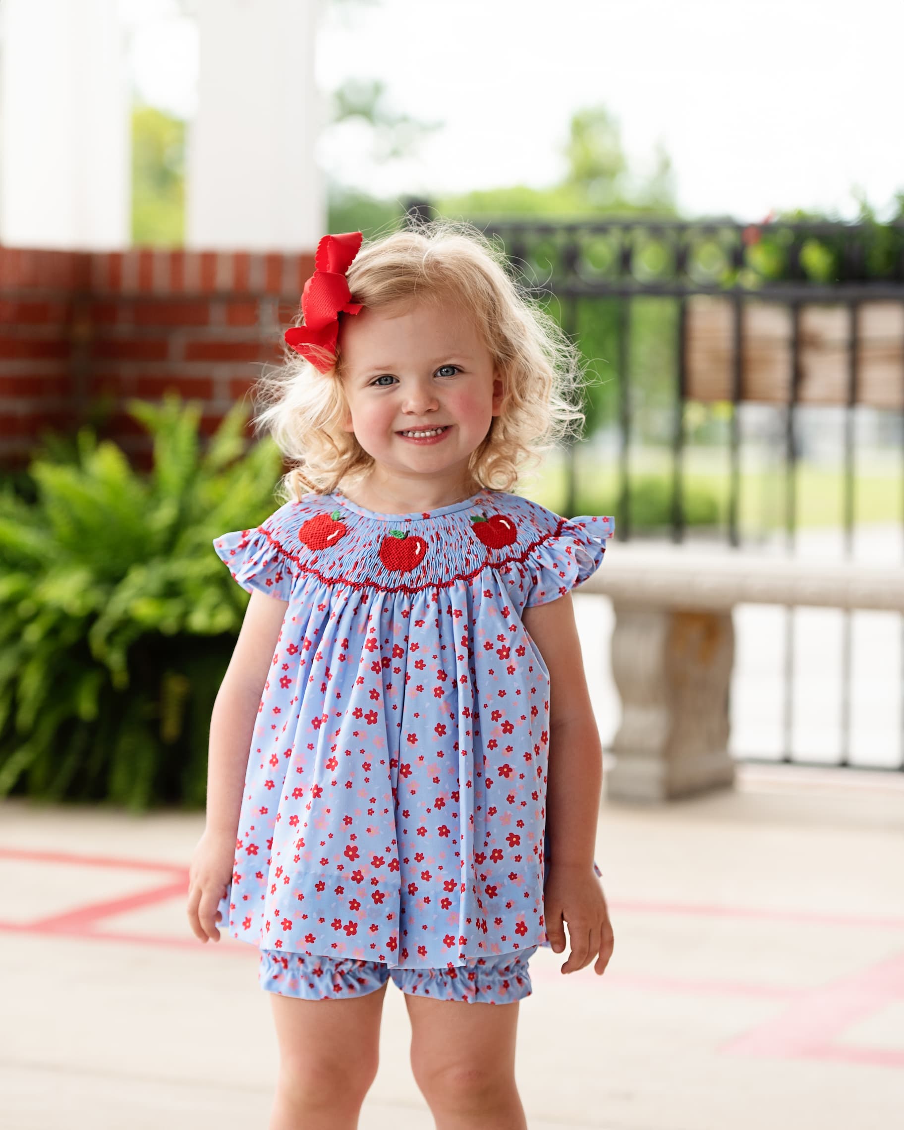 Child in floral outfit with smocked apples and a red bow.