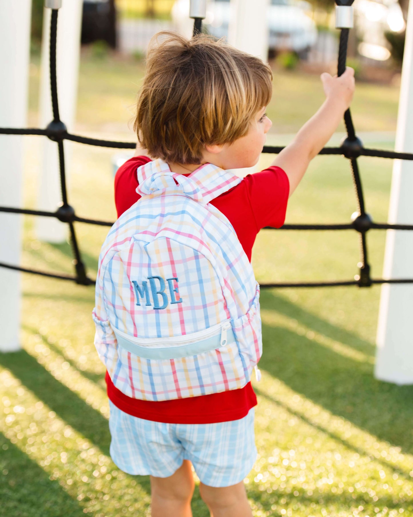 Child on a playground wearing a colorful plaid backpack with 'MBF' monogram.